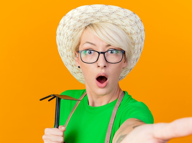 Foto gratuita mujer joven jardinero con pelo corto en delantal y sombrero sosteniendo azadón lookign en cámara siendo sorprendido de pie sobre fondo naranja