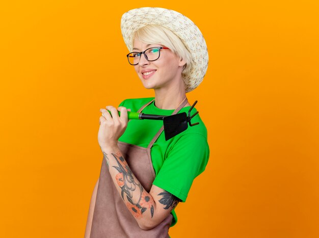 Mujer joven jardinero con pelo corto en delantal y sombrero sosteniendo azadón mirando a la cámara con una sonrisa en la cara de pie sobre fondo naranja