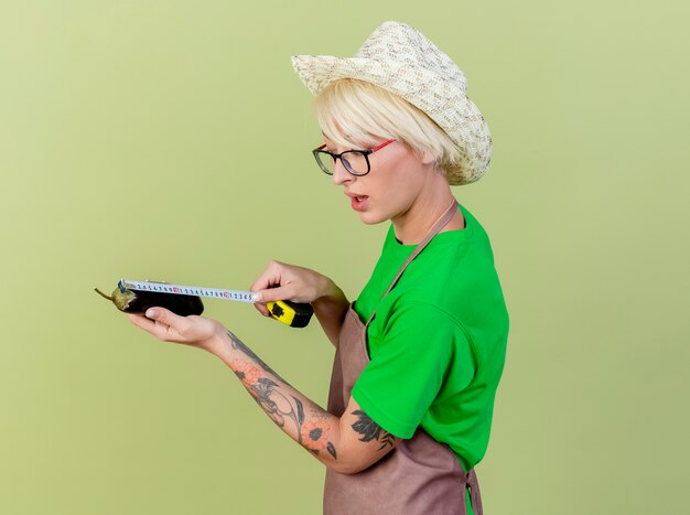Mujer joven jardinero con pelo corto en delantal y sombrero sosteniendo berenjena midiéndolo con cinta métrica mirando confiado de pie sobre fondo claro
