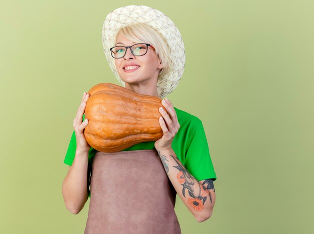 Mujer joven jardinero con pelo corto en delantal y sombrero sosteniendo calabaza mirando a la cámara con una sonrisa en la cara de pie sobre fondo claro