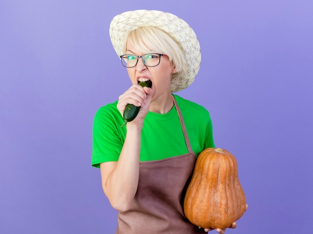 Foto gratuita mujer joven jardinero con pelo corto en delantal y sombrero sosteniendo calabaza morder pepino parado sobre fondo azul.