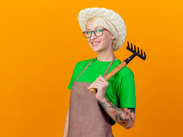 Mujer joven jardinero con pelo corto en delantal y sombrero sosteniendo mini rastrillo lookign a la cámara sonriendo alegremente de pie sobre fondo naranja