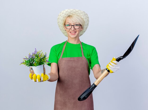 Mujer joven jardinero con pelo corto en delantal y sombrero sosteniendo pala y planta en maceta con sonrisa en la cara