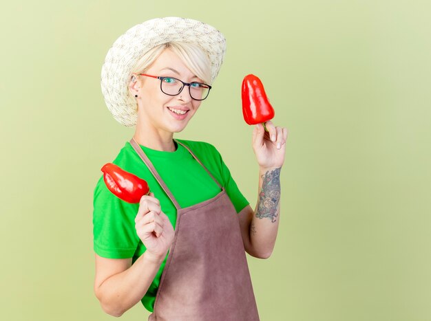 Mujer joven jardinero con pelo corto en delantal y sombrero sosteniendo pimientos rojos divirtiéndose sonriendo alegremente de pie sobre fondo claro