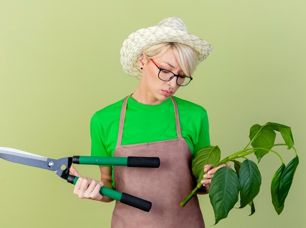 Mujer joven jardinero con pelo corto en delantal y sombrero sosteniendo planta y cortasetos mirando confundido e incierto de pie sobre fondo claro