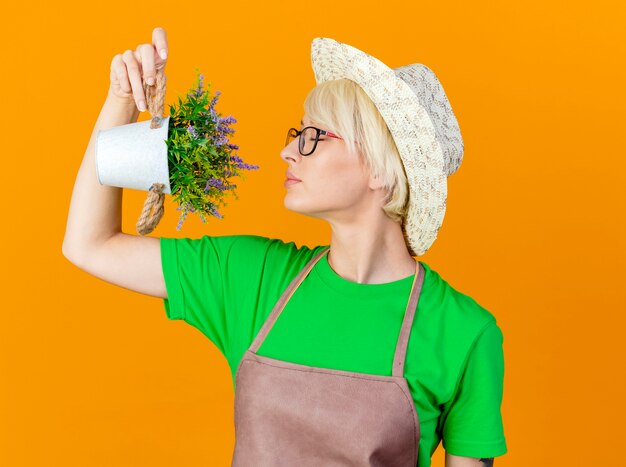 Mujer joven jardinero con pelo corto en delantal y sombrero sosteniendo planta en maceta