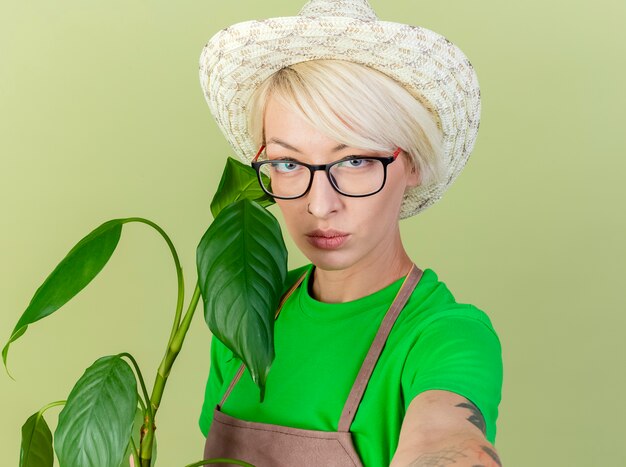 Mujer joven jardinero con pelo corto en delantal y sombrero sosteniendo planta