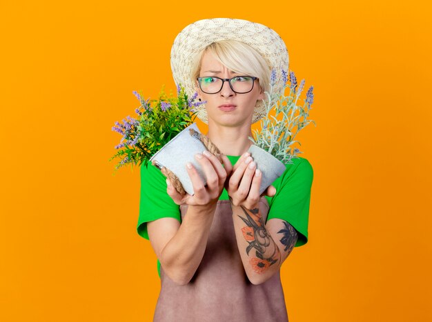 Mujer joven jardinero con pelo corto en delantal y sombrero sosteniendo plantas en macetas mirándolos confundido y disgustado de pie sobre fondo naranja