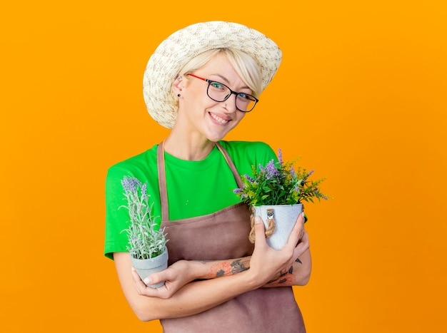 Mujer joven jardinero con pelo corto en delantal y sombrero sosteniendo plantas en macetas sonriendo con cara feliz mirando a la cámara de pie sobre fondo naranja