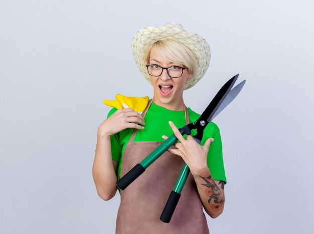 Mujer joven jardinero con pelo corto en delantal y sombrero sosteniendo podadoras de setos y guantes de goma sonriendo alegremente