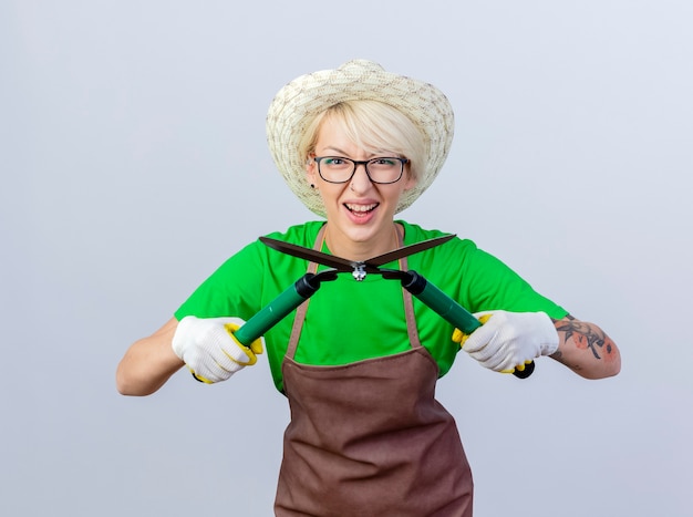 Mujer joven jardinero con pelo corto en delantal y sombrero sosteniendo podadoras de setos sonriendo alegremente