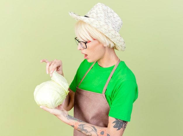 Foto gratuita mujer joven jardinero con pelo corto en delantal y sombrero sosteniendo repollo mirándolo intrigado parado sobre fondo claro