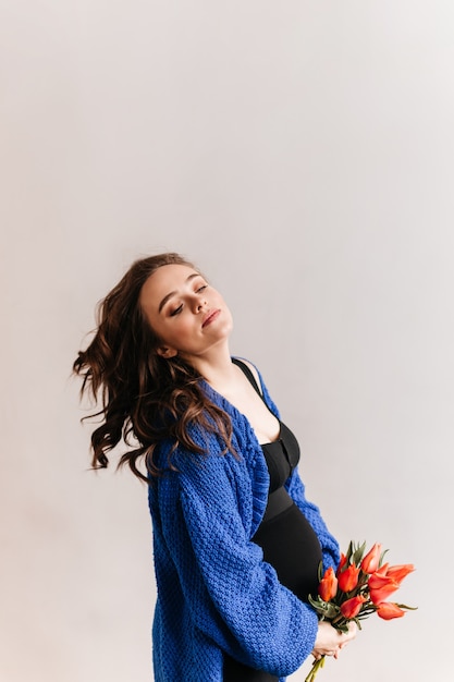Mujer joven juega cabello sobre fondo aislado. Mujer morena embarazada en cardigan azul tiene ramo de tulipanes.