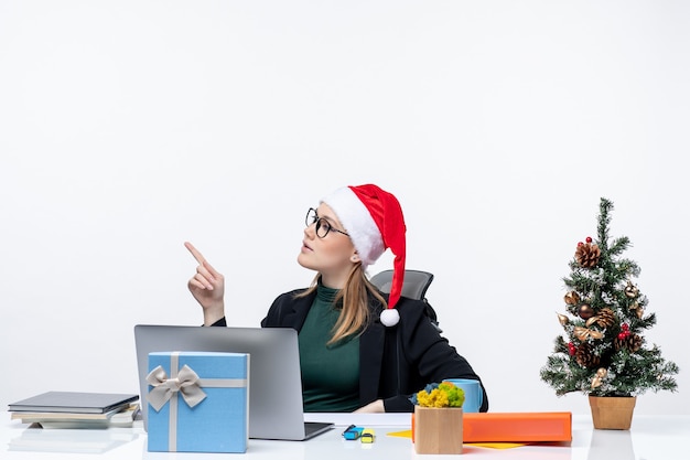Mujer joven jugando con sombrero de santa claus y anteojos sentado en una mesa con un árbol de Navidad y un regalo