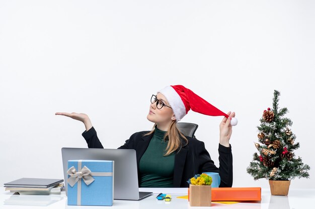 Mujer joven jugando con sombrero de santa claus y anteojos sentado en una mesa con un árbol de Navidad y un regalo