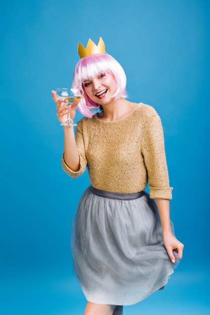 Mujer joven juguetona divertida con el pelo rosado cortado, champán expresando positividad. Corona dorada en la cabeza, falda de tul gris, vítores, gran fiesta de cumpleaños, emociones positivas en la cara.