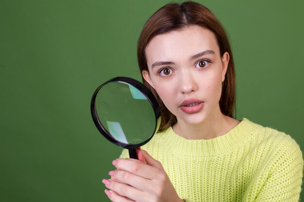 Mujer joven con labios grandes marrones de maquillaje natural perfecto en suéter casual en pared verde con lupa buscando