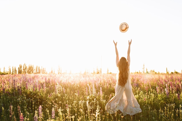 La mujer joven lanza para arriba el sombrero de paja en campo de flor en puesta del sol.
