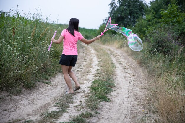 Una mujer joven lanza enormes pompas de jabón en la hermosa naturaleza de fondo, vista posterior.