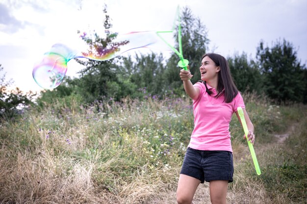 Una mujer joven lanza grandes pompas de jabón de colores entre la hierba en la naturaleza.