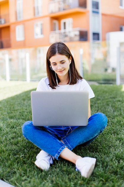 Mujer joven con laptop sentada sobre la hierba verde y mirando a una pantalla al aire libre