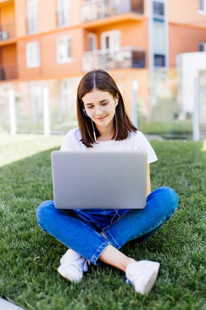 Mujer joven con laptop sentada sobre la hierba verde y mirando a una pantalla al aire libre