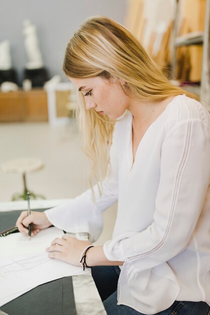 Mujer joven lateral que bosqueja en el papel con el lápiz en el banco de trabajo