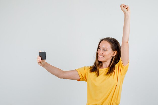 Mujer joven levantando el brazo mientras toma selfie en camiseta, pantalones cortos y se ve alegre. vista frontal.