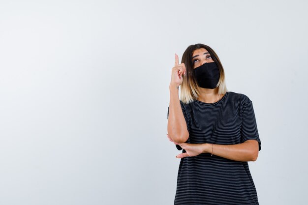 Mujer joven levantando el dedo índice en gesto de eureka, sosteniendo la mano debajo del codo en vestido negro, máscara negra y mirando sensible, vista frontal.