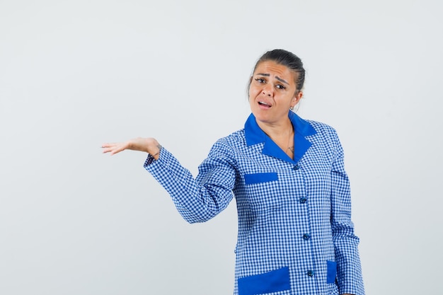 Mujer joven levantando la mano como sosteniendo algo en la camisa de pijama de cuadros azules y luciendo bonita. vista frontal.