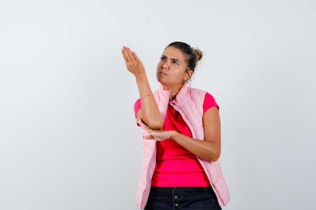 Mujer joven levantando la mano por encima, sosteniendo una mano en el codo en camiseta rosa y chaqueta y mirando enfocado