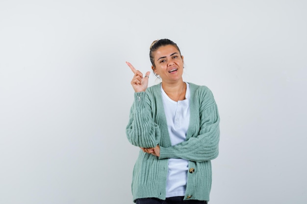 Mujer joven levantando una mano en gesto eureka mientras sostiene la mano en el codo en camiseta blanca y chaqueta de punto verde menta y parece feliz