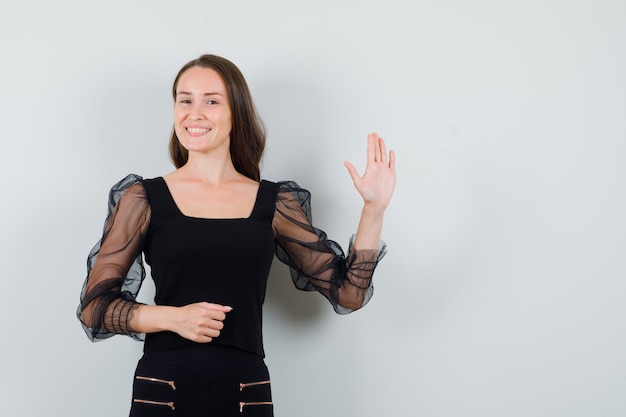 Mujer joven levantando la mano para saludar en blusa negra y mirando complacido