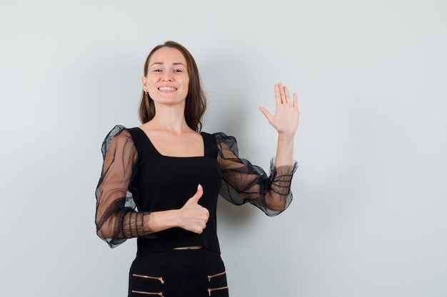 Mujer joven levantando la mano para saludar mientras muestra el pulgar hacia arriba en una blusa negra y se ve alegre. vista frontal.