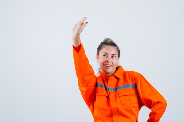 Mujer joven levantando la mano para saludos en uniforme de trabajador y mirando contento, vista frontal.