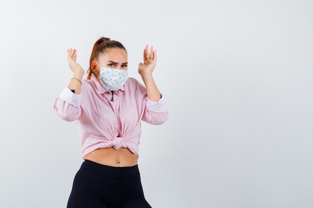 Foto gratuita mujer joven levantando las manos cerca de la cabeza en camisa, pantalones, máscara médica y luciendo estresada. vista frontal.