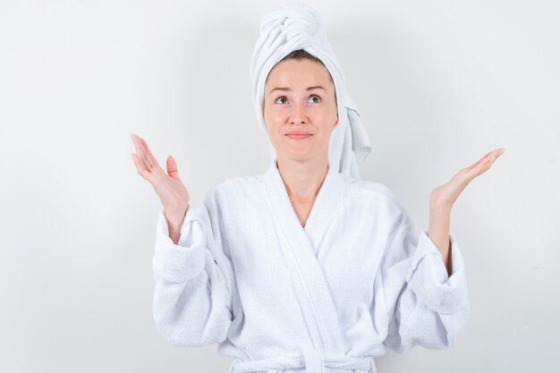 Mujer joven levantando las palmas de las manos mientras mira hacia arriba en bata de baño blanca, toalla y parece esperanzada. vista frontal.