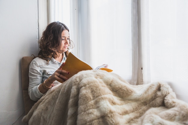 Mujer joven leyendo un libro junto a la ventana