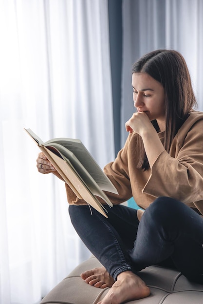 Foto gratuita una mujer joven está leyendo un libro sentada en un sillón junto a la ventana.