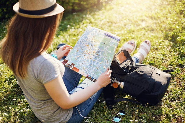 Mujer joven leyendo el mapa de la ciudad sentada en el parque con sombrero y mochila Concepto de viaje para jóvenes