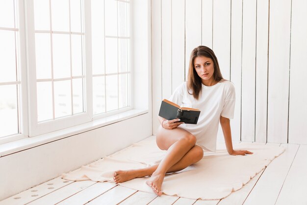 Mujer joven con el libro en manta junto a la ventana