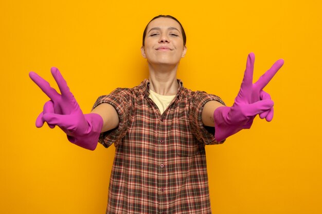 Mujer joven de la limpieza en ropa casual en guantes de goma que muestra el signo v feliz y positivo