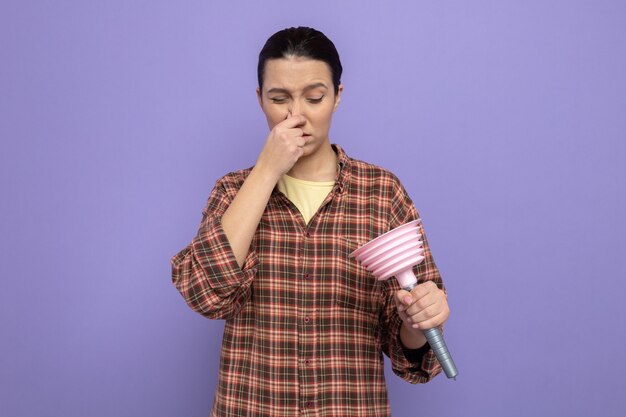Mujer joven de la limpieza en ropa casual sosteniendo el émbolo mirándola cerrando la nariz con los dedos que sufren de hedor de pie sobre la pared púrpura