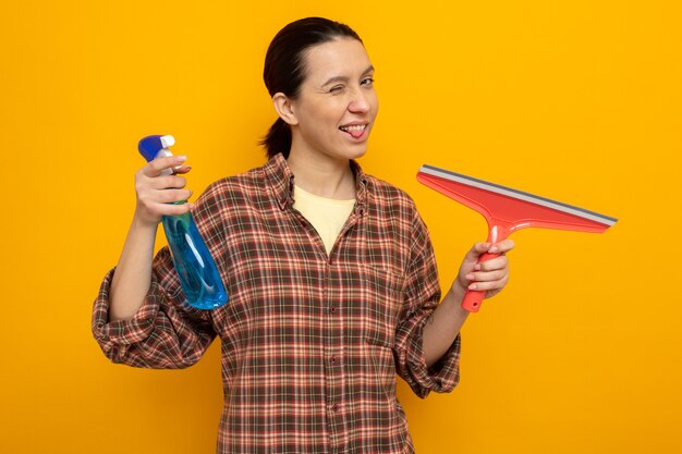 Mujer joven de la limpieza en ropa casual con spray de limpieza y un trapeador mirando al frente feliz y positivo que sobresale la lengua de pie sobre la pared naranja