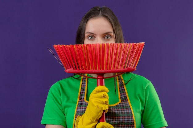 Mujer joven de limpieza vistiendo uniforme en guantes con la cara cubierta con un trapeador en la pared púrpura