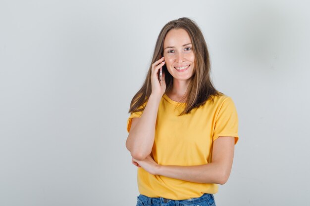 Mujer joven llamando a alguien por teléfono móvil en camiseta amarilla y mirando alegre