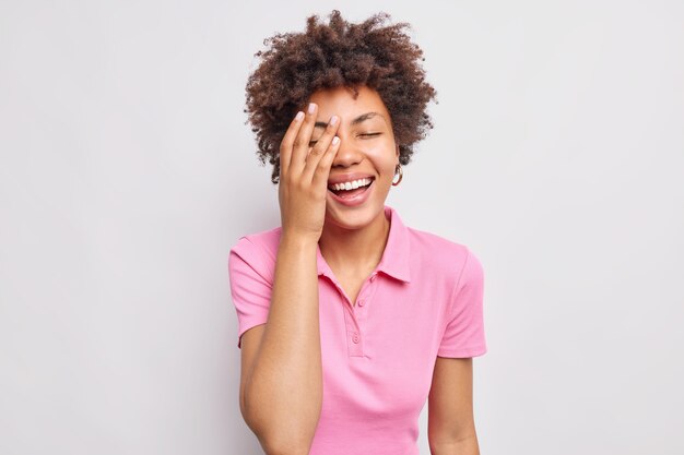 La mujer joven llena de alegría hace que la palma de la cara se ría positivamente se siente feliz de no esconder sus emociones positivas y auténticas vestidas casualmente aisladas en la pared blanca