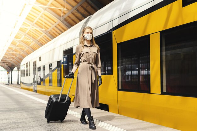 Mujer joven con una maleta con guantes y mascarilla caminando en una estación de tren - COVID-19