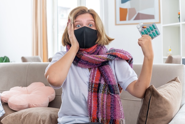 Mujer joven malsana con bufanda caliente alrededor del cuello con máscara protectora facial que se siente mal y enfermo que sufre de gripe y píldoras de retención de frío con aspecto confundido sentado en el sofá en la sala de estar luminosa