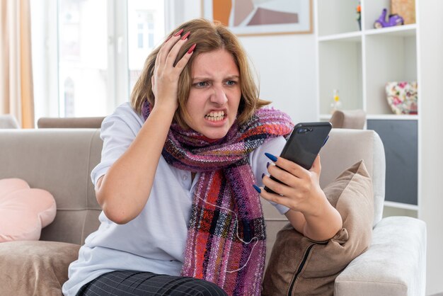 Mujer joven malsana con bufanda caliente alrededor del cuello que se siente mal y enferma que sufre de gripe y frío mirando el teléfono inteligente enojado sentado en el sofá en la sala de estar iluminada